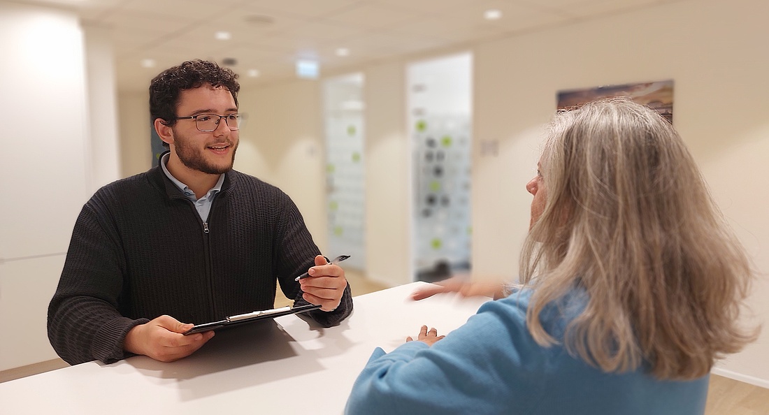 Foto: Ein junger Mann mit dunklen Locken und Brille mit Mappe in der Hand steht an einem Empfangspult und spricht mit einer Frau mit grauen langen Haaren. 