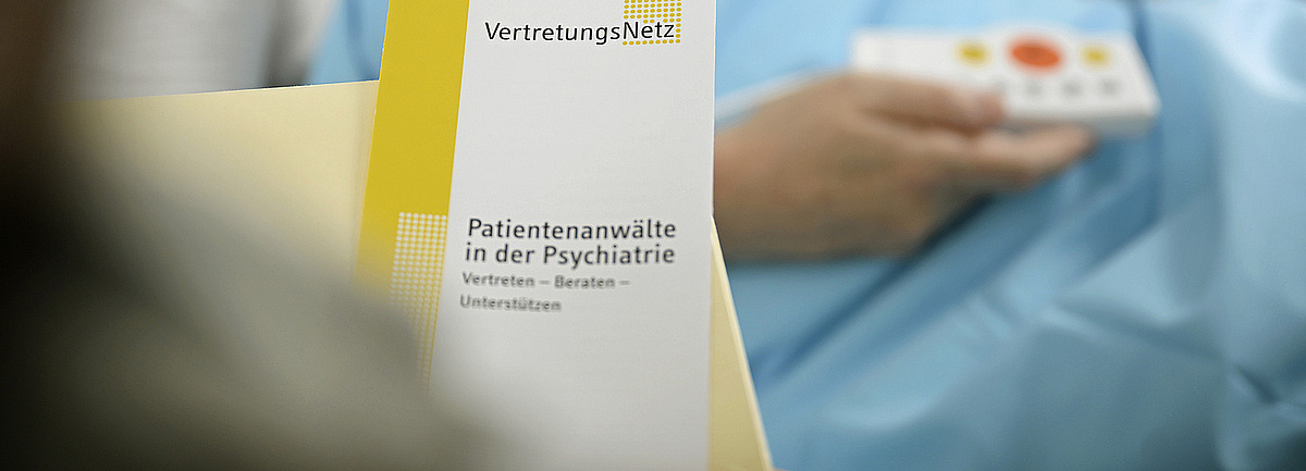 Der Folder der Patientenanwaltschaft im Vordergrund, im Hintergrund eine im Krankenbett liegende Person, die ein Rufhilfegerät hält. 