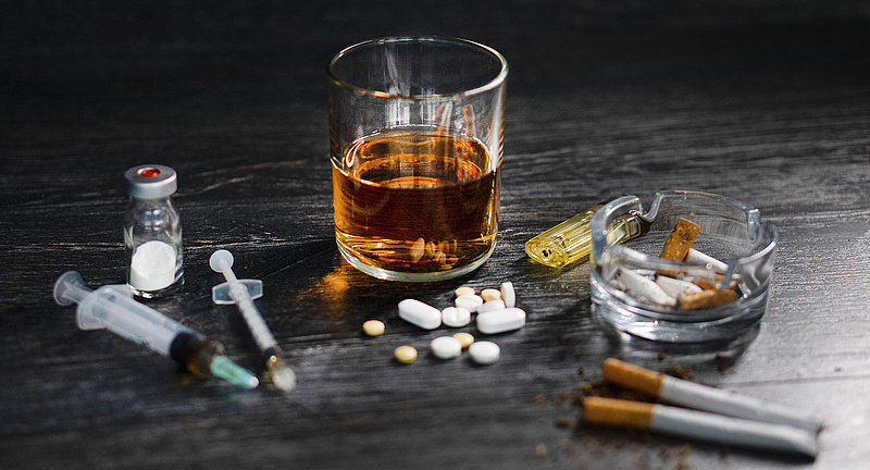 Suchtmittel auf einem dunklen Tisch: Zigaretten, ein Glas Whiskey, Tabletten und Drogen
