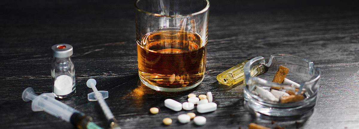 Suchtmittel auf einem dunklen Tisch: Zigaretten, ein Glas Whiskey, Tabletten und Drogen