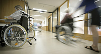 Foto: Auf dem Foto sieht man einen laufenden Betrieb in einem Pflegeheim. Ein Mann in einem Rollstuhl ist links am Foto zu sehen und rechts wird eine andere Person im Rollstuhl vorbeigefahren. Sie befinden sich in einem Flur. 