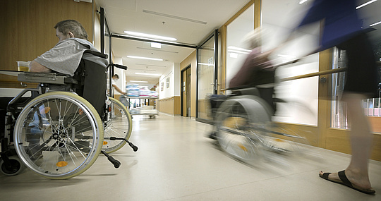 Foto: Auf dem Foto sieht man einen laufenden Betrieb in einem Pflegeheim. Ein Mann in einem Rollstuhl ist links am Foto zu sehen und rechts wird eine andere Person im Rollstuhl vorbeigefahren. Sie befinden sich in einem Flur. 