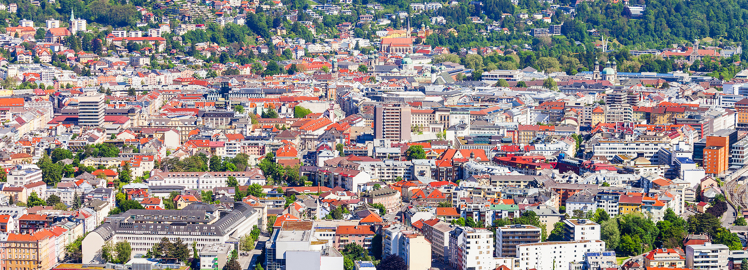 Erwachsenenvertretung in Innsbruck, VertretungsNetz. FC: iStock Foto von Innsbruck: Luftaufnahme einer österreichischen Stadt bei sonnigem Wetter mit dicht bebauten Wohn- und Geschäftshäusern, einer gelben Kirche mit Turm im Vordergrund, Straßen mit Verkehr, einem Bahnhofs- und Industrieareal sowie grünen Hügeln im Hintergrund.