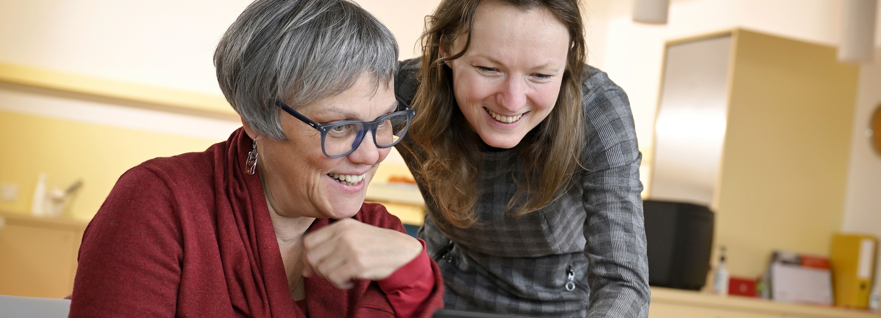 Foto: Zwei Frauen schauen lächelnd auf einen Laptop-Bildschirm. Eine Frau sitzt und tippt auf dem Laptop, während die andere Frau dahinter steht und sich über den Bildschirm beugt.