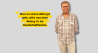 Ein Herr mit braunen Haaren und Brille, karriertem Hemd und beiger Hose steht vor einem grauen Hintergrund und lächelt in die Kamera. Links daneben steht gelb unterstrichen "Wenn es einem selbst gut geht, sollte man einen Beitrag für die Gesellschaft leisten.  