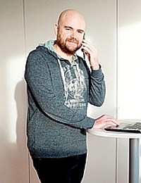 Mann mit Bart in dunkelgrauem Hoodie steht an einem Stehtisch, telefoniert und nutzt einen Laptop in einem hellen Innenraum.