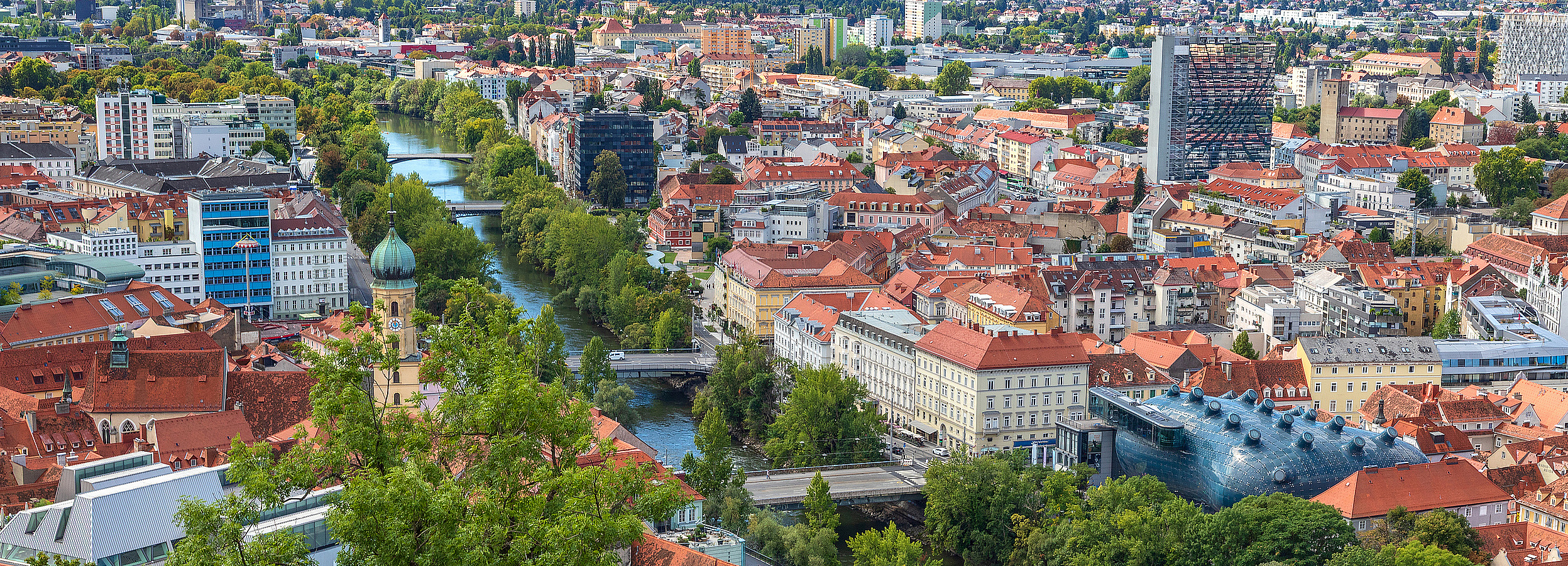 Erwachsenenvertretung in Graz, VertretungsNetz. FC: iStock Foto von Graz: Panoramablick von oben auf eine Stadt mit roten Ziegeldächern, historischem Uhrturm und modernen Gebäuden, einem Fluss mit mehreren Brücken, vielen Bäumen und Grünflächen sowie sanften Hügeln und Bergen am Horizont unter leicht bewölktem Himmel.