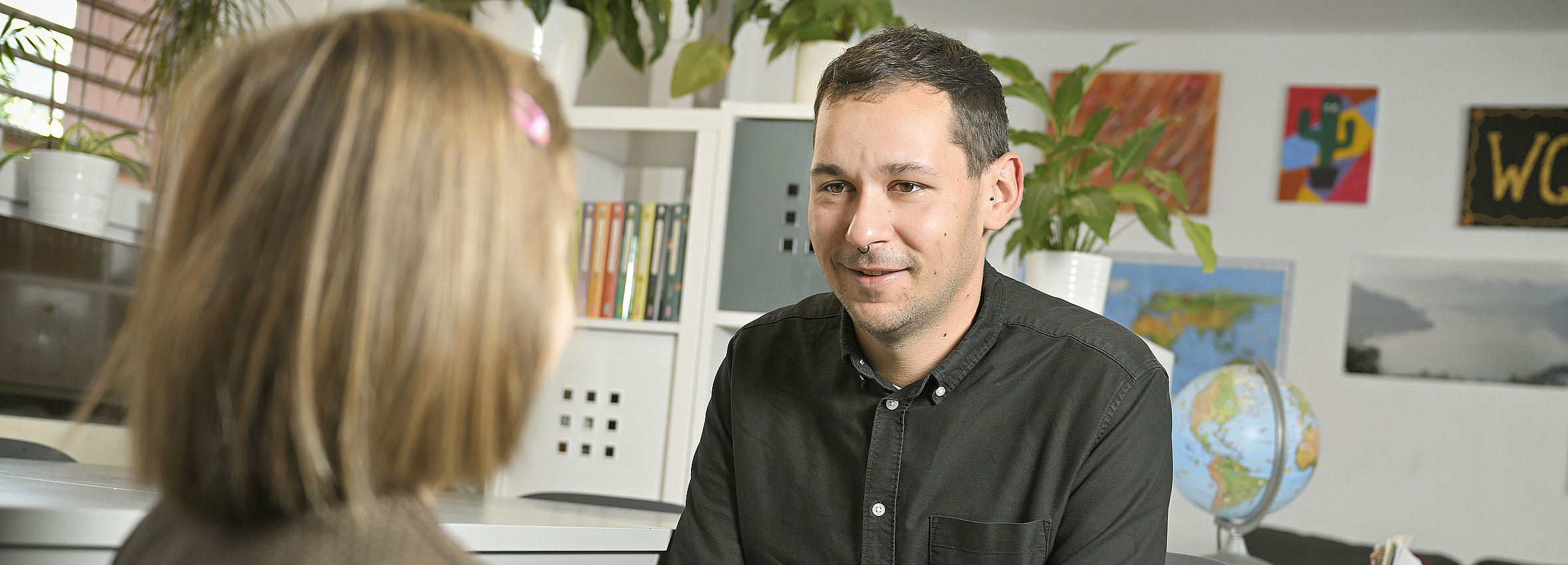 Foto: Kind sitzt mit Rücken zur Kamera. Ihm gegenüber sitzt ein junger Mann mit dunkelbraunen Haaren und lächelt es an. 