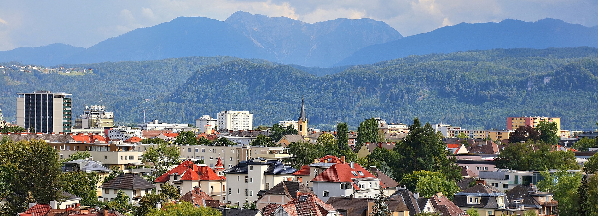 Erwachsenenvertretung in Klagenfurt, VertretungsNetz. FC: iStock Foto von Klagenfurt: Städtische Ansicht mit vielfältiger Wohnbebauung, Kirchturm und modernen Hochhäusern vor einer Kulisse aus bewaldeten Hügeln und hohen Bergen unter teils bewölktem Himmel.