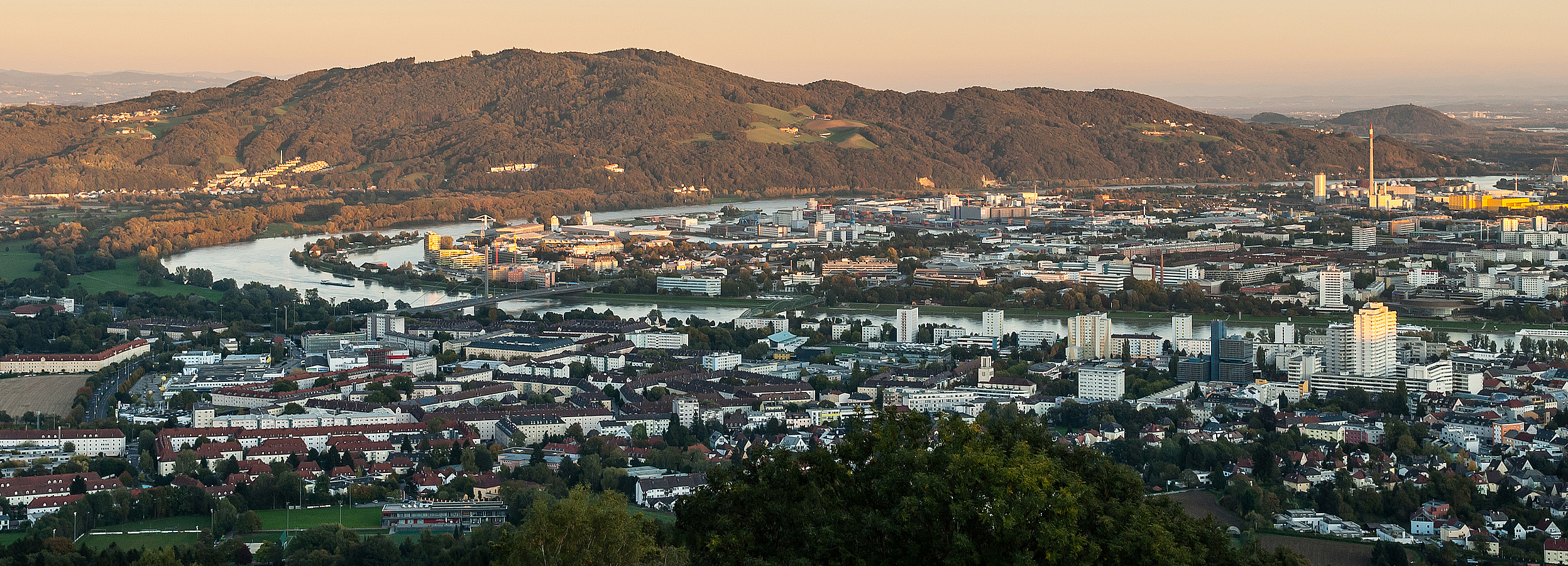 Foto von Linz: Panoramablick über eine Stadt an einem geschwungenen Fluss, umgeben von bewaldeten Hügeln und grünen Flächen, aufgenommen bei warmem Licht in der Dämmerung.