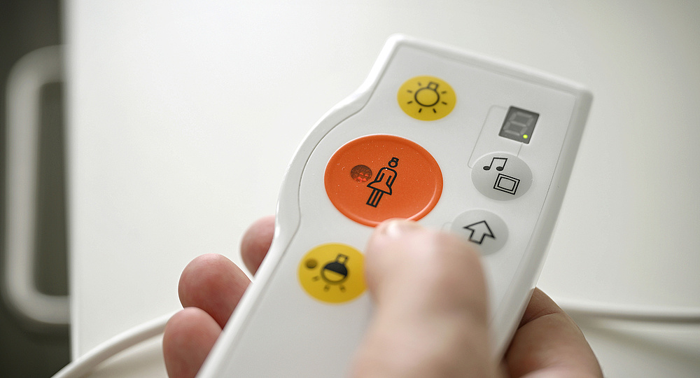 Personalmangel in der Psychiatrie. Am Bild ist eine Hand zu sehen, die eine Notruftaste vom Krankenhaus hält. Auf der Fernbedienung ist in der Mitte ein roter Button mit einer Grafik von einer Krankenschwester zu sehen.