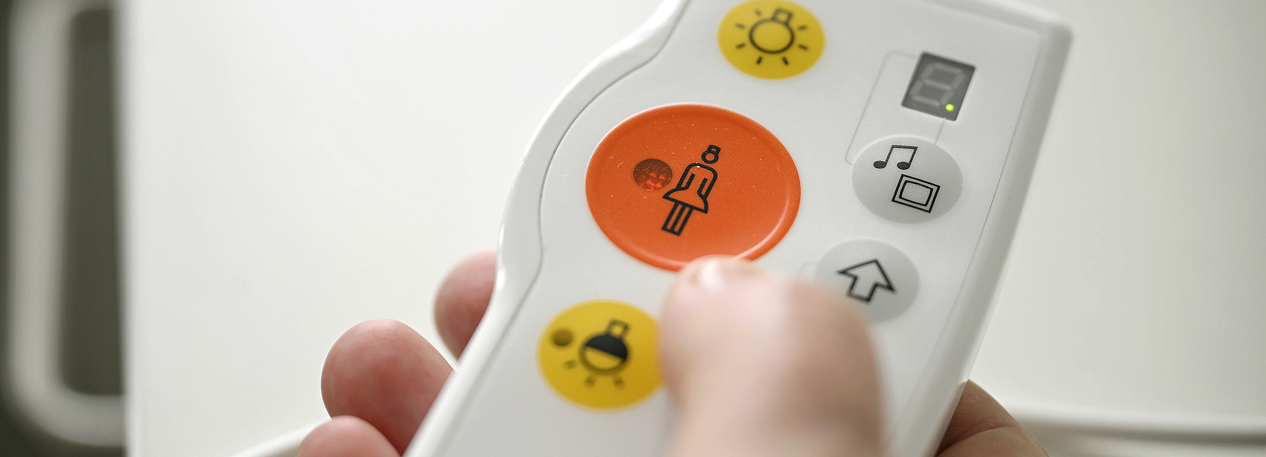 Auf dem Bild ist eine Hand zu sehen, die eine Notruftaste eines Krankenhauses hält. In der Mitte der Fernbedienung ist ein roter Button mit einer Grafik einer Krankenschwester. 