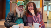 Foto: Ein Mann und eine Frau sitzen an einem Tisch nebeneinander, vor ihnen zwei Kaffetassen. Sie lächeln in die Kamer. Gottfried und Lena.