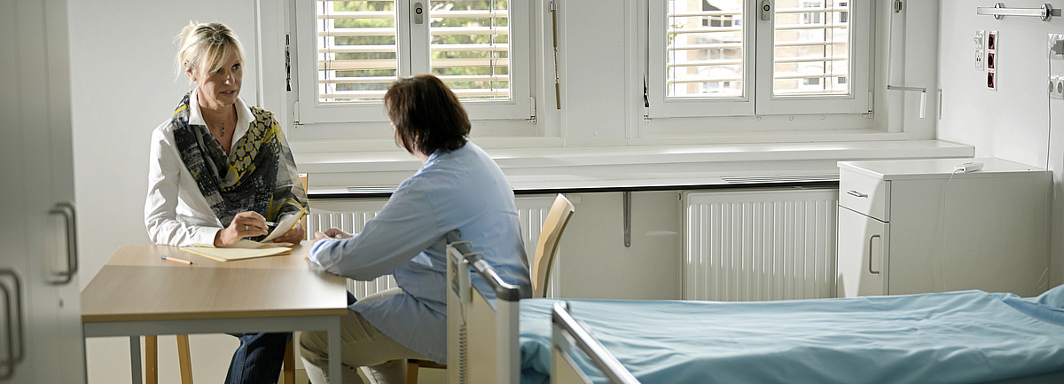 Patientenanwältin im Gespräch mit einer Patientin an eine Tisch im Krankenhauszimmer, daneben ein Bett