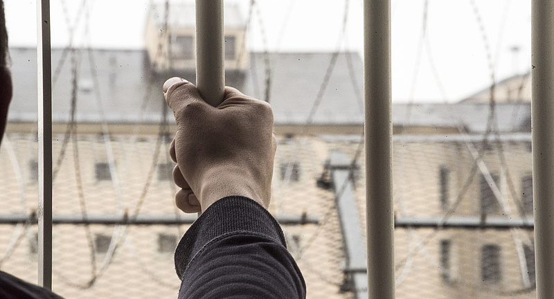 Foto: Hand eines Häftlings ergreift einen Stab des vergitterten Gefängisses, hinter den Gitterstäben erkennt man einen weiteren Gefängnistrakt