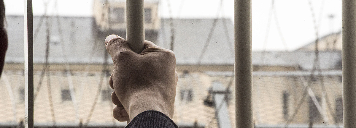Foto: Hand eines Häftlings ergreift einen Stab des vergitterten Gefängisses, vor den Gitterstäben erkennt man einen weiteren Gefängnistrakt