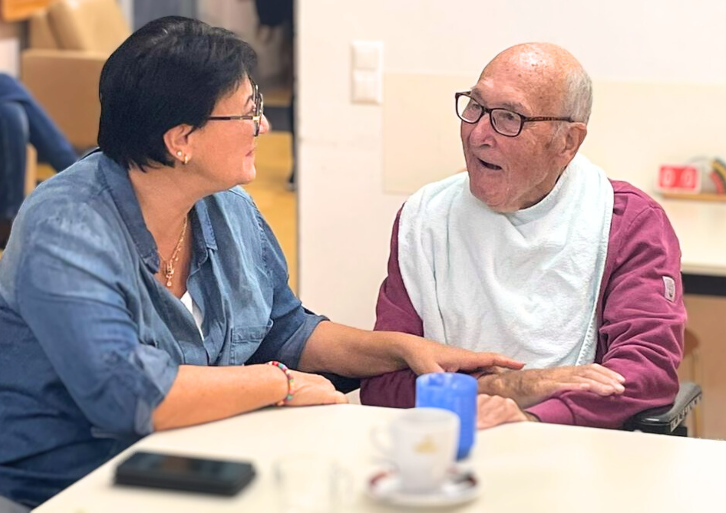 Foto: Eine Frau sitzt mit einem älteren Herren an einem Tisch, sie lächeln sich an.