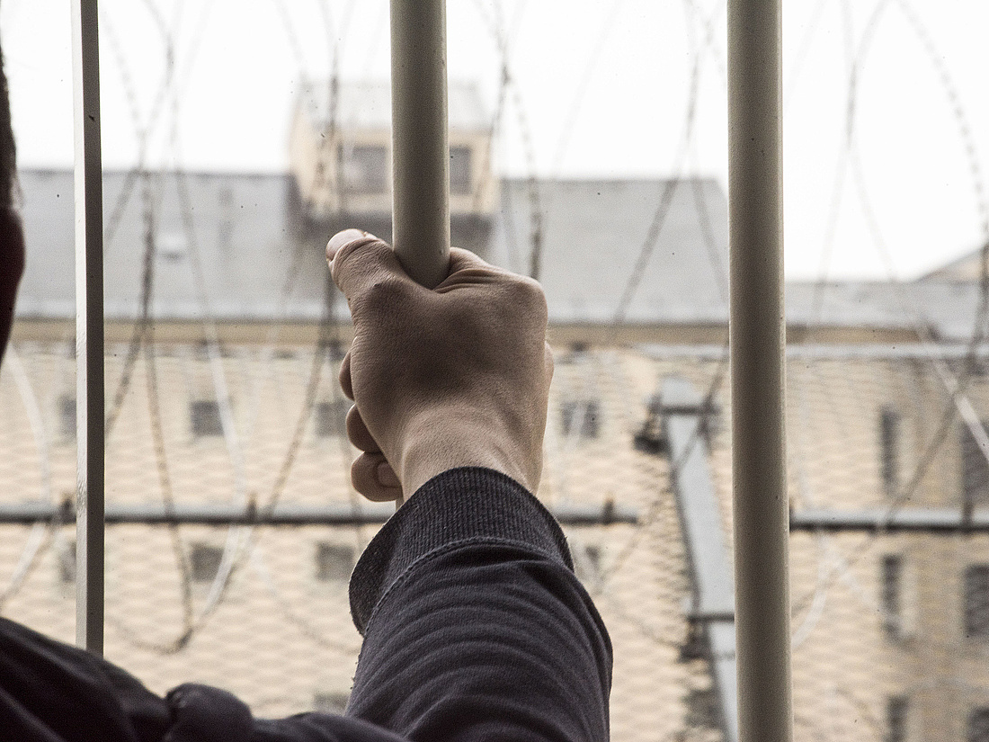 Foto: die Hand eines Gefangenen auf Gitterstäben, Aussicht auf Gefängnisbau