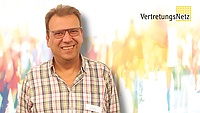 Porträt einer lächelnden Person mit Brille und kariertem Hemd, mit Namensschild, vor hellem unscharfem Hintergrund; oben rechts das Logo „VertretungsNetz“