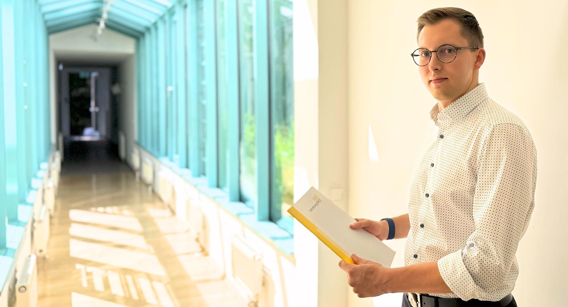Foto: Junger Mann mit runder Brille und hell gepunktetem Hemd in einem lichtdurchfluteten Glasgang; er hält eine gelbe Mappe und blickt in die Kamera.