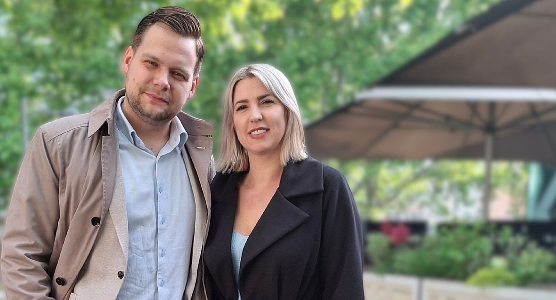 Foto: Ein Mann mit kurz geschnittenem braunem Haar, hellblauem Hemd und beigem Trenchcoat und eine Frau mit schulterlangem blondem Haar und schwarzem Mantel stehen lächelnd nebeneinander im Freien vor grünen Bäumen und einem großen Sonnenschirm.