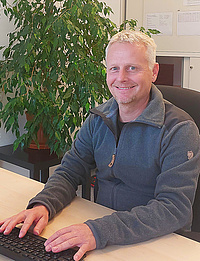 Grauhaariger Mann in dunkelblauer Fleecejacke sitzt lächelnd an einem Schreibtisch und tippt auf einer Tastatur; im Hintergrund eine große grüne Zimmerpflanze im Büro.