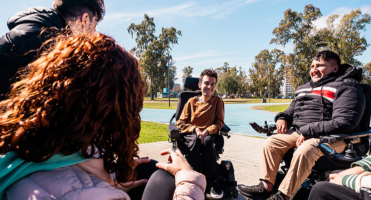 nf Menschen unterhalten sich lachend im sonnigen Park; zwei Personen in elektrischen Rollstühlen, mit blauer Laufbahn und Bäumen im Hintergrund.