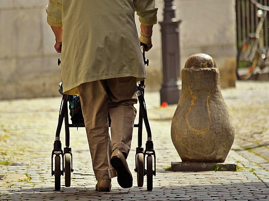 Foto einer Frau, Rückenansicht, die mit Hilfe eines Rollators geht