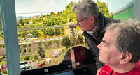 Zwei ältere Menschen beugen sich in einer Ausstellung über eine detailreiche Miniaturlandschaft mit Modellbahn, Fahrzeugen und bewaldeten Hügeln.