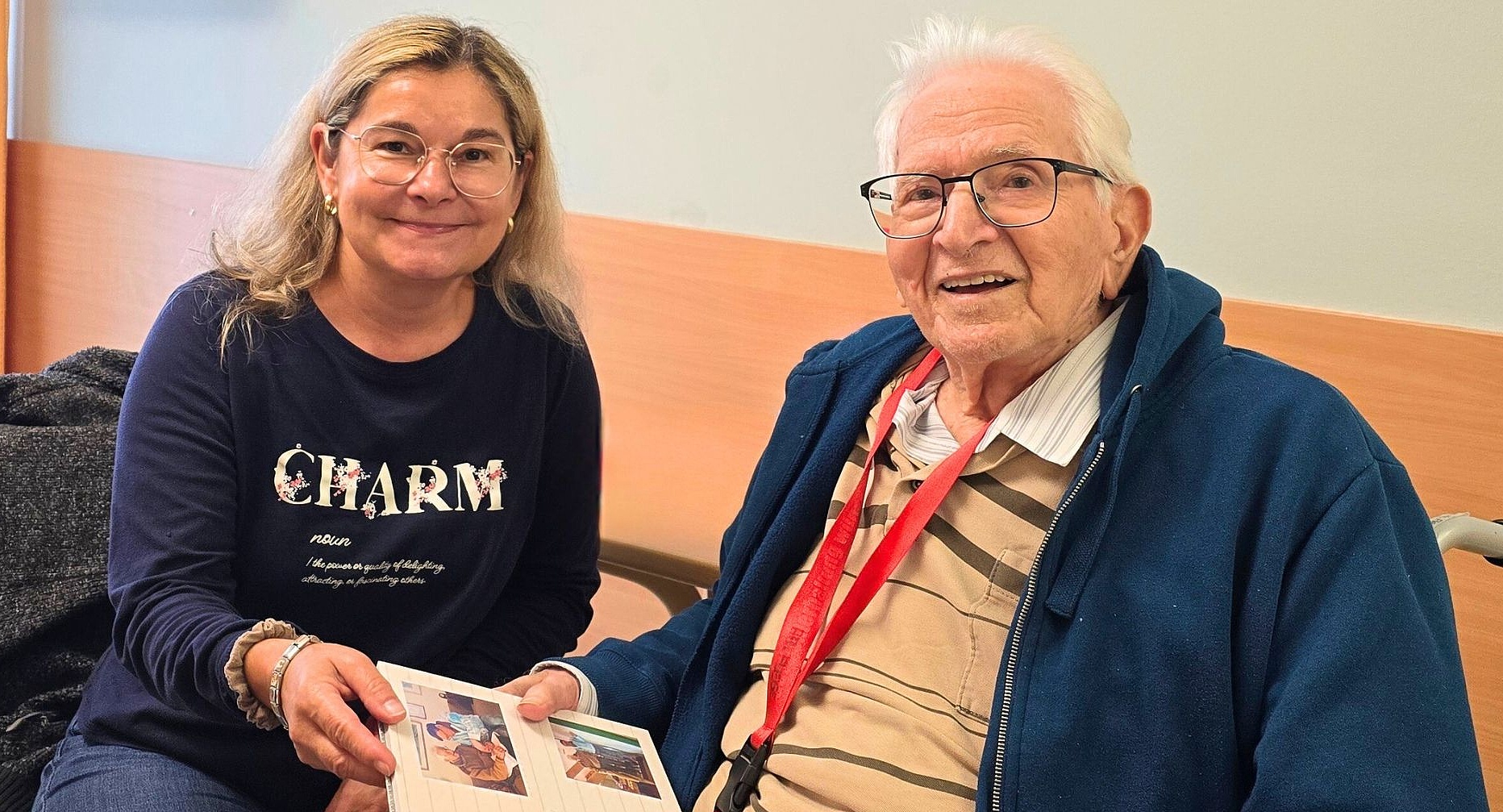 Foto: Eine lächelnde Frau sitzt neben einem älteren Mann in einer Pflegeeinrichtung; beide tragen Brillen und halten ein Fotoalbum, während sie freundlich in die Kamera blicken.