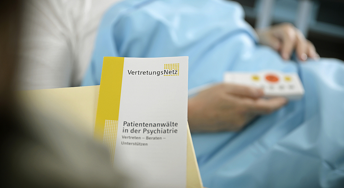 Oberkörper einer Patientin im Krankenhaus-Bett, Folder Patientenanwält:innen wird überreicht