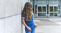 Foto: jugendliches Mädchen lehnt an einer Mauer vor dem Krankenhaus, das Gesicht abgewendet von der Kamera, sieht sie in Richtung Türen