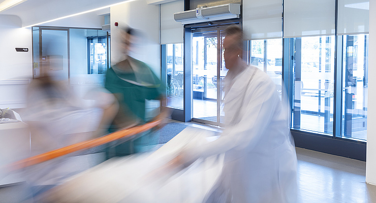 Ärzte und Pfleger:innen schieben ein Bett mit einem Patienten. Dahinter Glasfronten eines Krankenhaus-Foyers