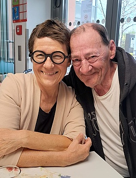 Foto: Zwei Menschen sitzen sich an einem Tisch nebeneinander und lachen in die Kamera. Eine gelbe Blume in einer grünen Vase und Getränke stehen auf dem Tisch.