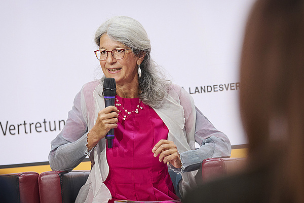 Susanne Jaquemar sitzend, sie hat lange graue Haare und eine Brille, sie trägt ein magentafarbenes Kleid und eine silberne Weste