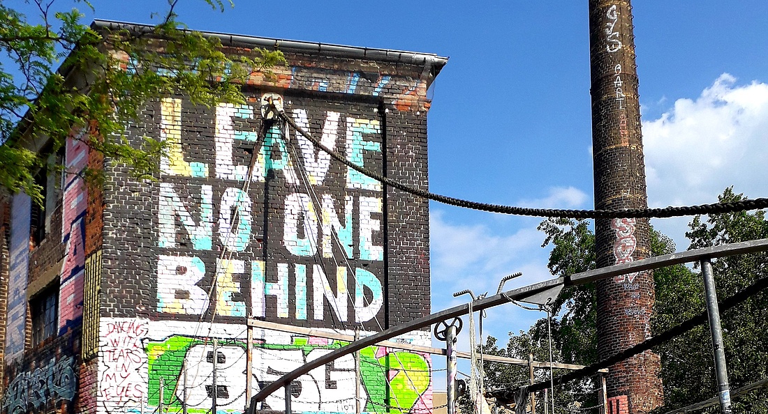 Foto einer alten Fabrik, auf den Mauern ein buntes Graffiti mit dem Text "Leave no one behind"