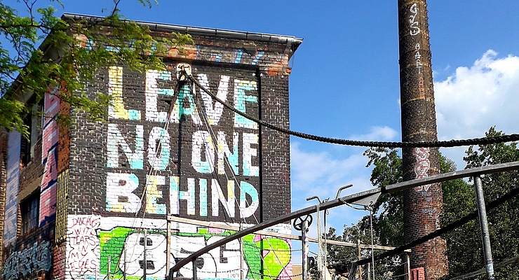 Foto einer alten Fabrik, auf den Mauern ein buntes Graffiti mit dem Text "Leave no one behind"