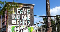 Foto einer alten Fabrik, auf den Mauern ein buntes Graffiti mit dem Text "Leave no one behind"