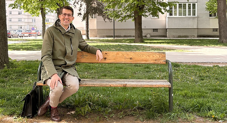 Foto: Lächelnder Mann mit Brille sitzt auf einer Holzbank in einem Park; neben der Bank steht eine schwarze Tasche.