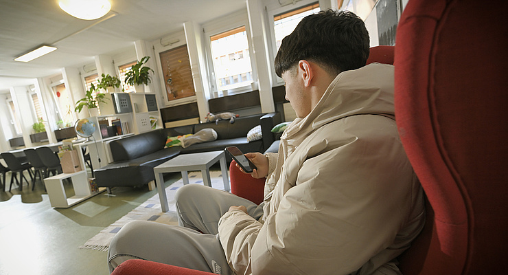 ein Jugendlicher mit schwarzen kurzen Haaren und beiger Jacke sitzt im Aufenthaltsraum einer WG auf einem roten Polsterstuhl und sieht auf sein Handy