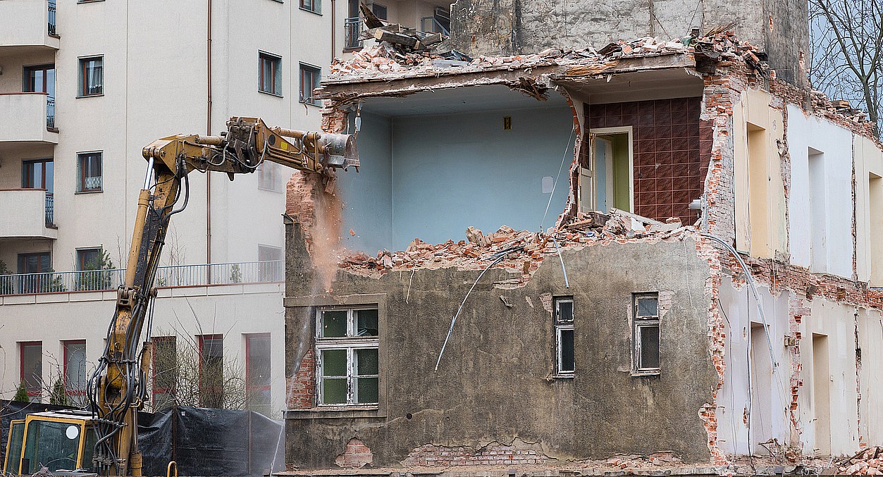 ein oranger Bagger reißt soeben ein Haus ein, es steht nur noch die Hälfte, man sieht in ein offenes blaugestrichenes Zimmer