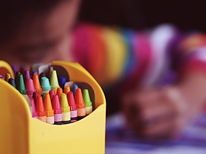 Schachtel mit Farbstiften, im Hintergrund ist die Hand eines malenden Kindes erkennbar.