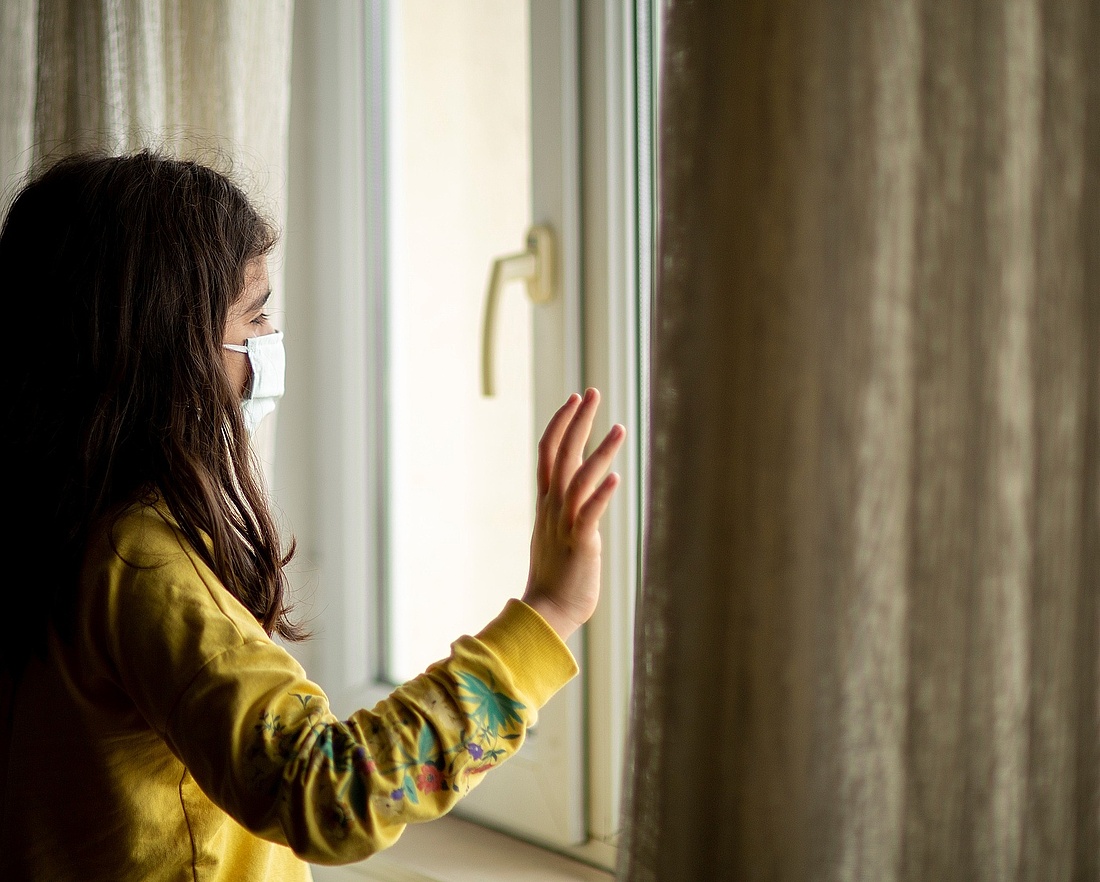 Foto: Junges Mädchen mit Maske schaut aus verschlossenem Fenster hinaus