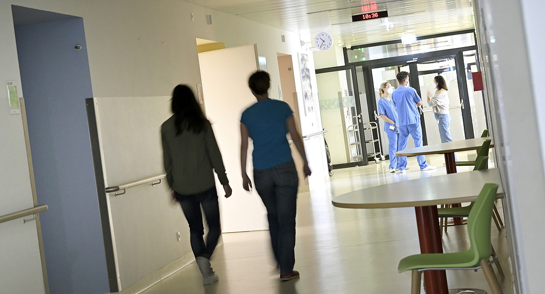 Ein Krankenhausflur, Rückenansicht von zwei gehenden Frauen, im Hintergrund stehen zwei Pflegekräfte in blauer Arbeitskleidung und sprechen mit einer Besucherin