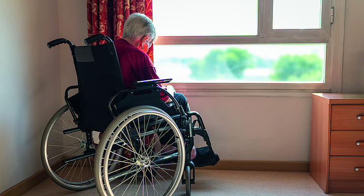 eine alte Frau sitzt im Rollstuhl in ihrem Pflegeheimzimmer und sieht aus dem Fenster. Sie wirkt traurig