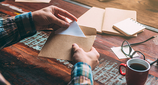 Foto: Eine Hand zieht einen Brief aus einem braunen Papierumschlag. Der Schreibtisch ist mit Notizbüchern, einer Kaffeetasse, einer Brille und weiteren Materialien bestückt.