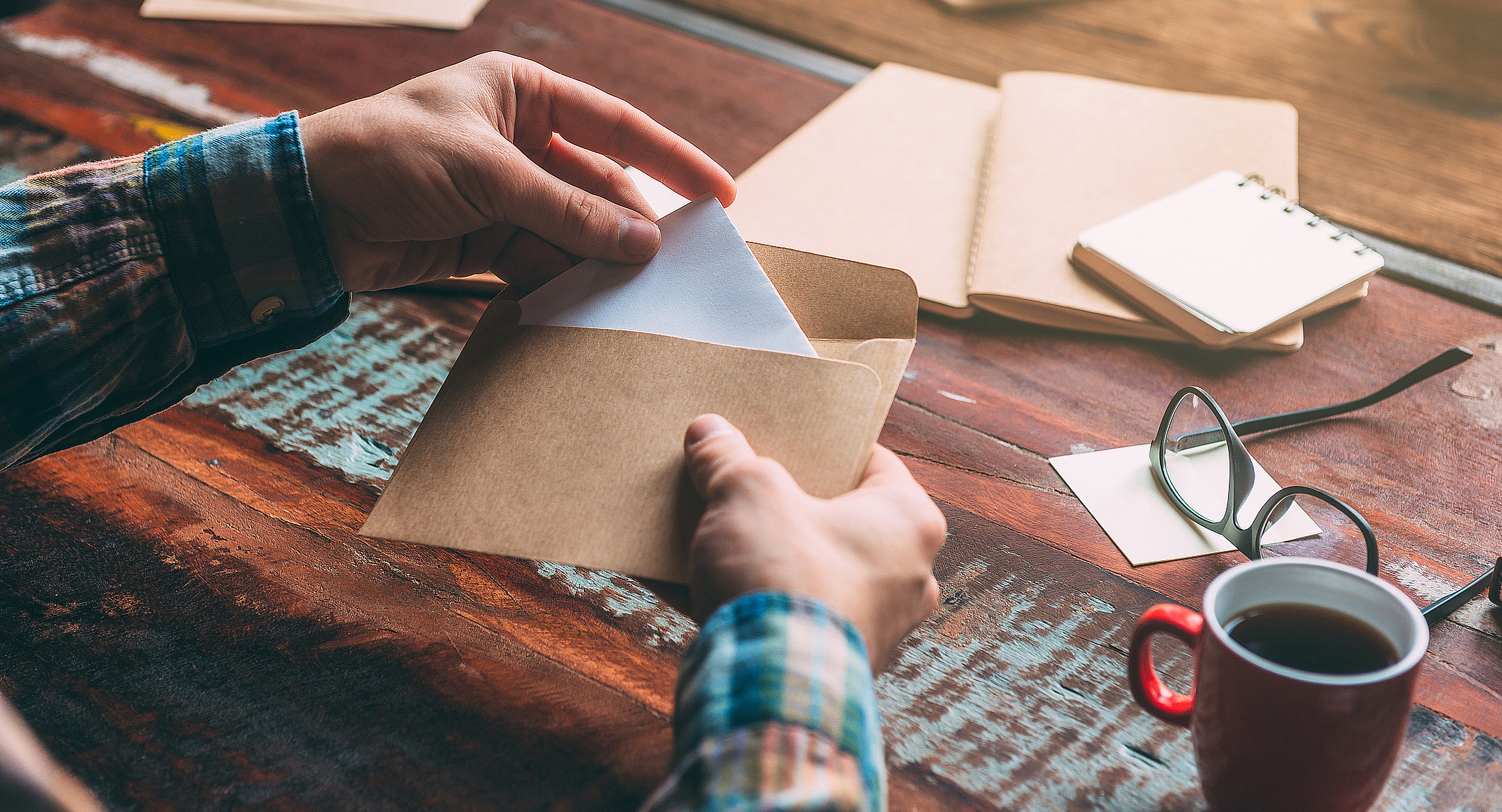 Foto: Eine Hand zieht einen Brief aus einem braunen Papierumschlag. Der Schreibtisch ist mit Notizbüchern, einer Kaffeetasse, einer Brille und weiteren Materialien bestückt.