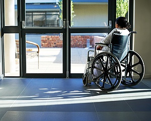 Foto von Frau im Rollstuhl im Pflegeheim, sieht durch geschlossene Tür