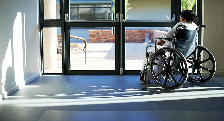 eine Frau im Rollstuhl sitzt vor einer geschlossenen Glastür und blickt ins Freie