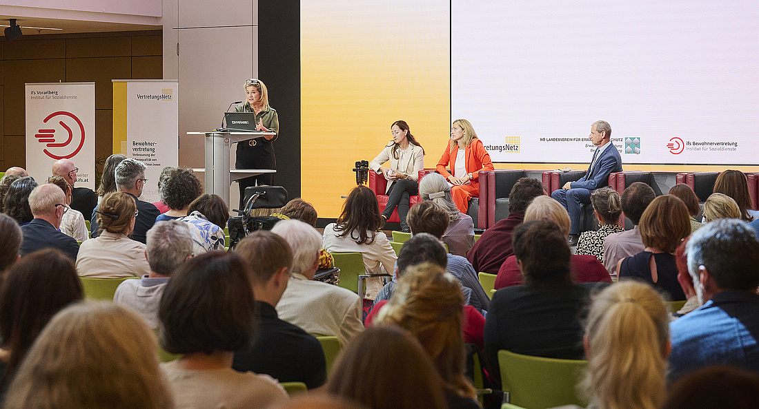 Grainne Nebois am Stehpult, sie trägt blonde lange Haare und eine olivgrüne Bluse. Vor ihr Publikum in Rückenansicht. 